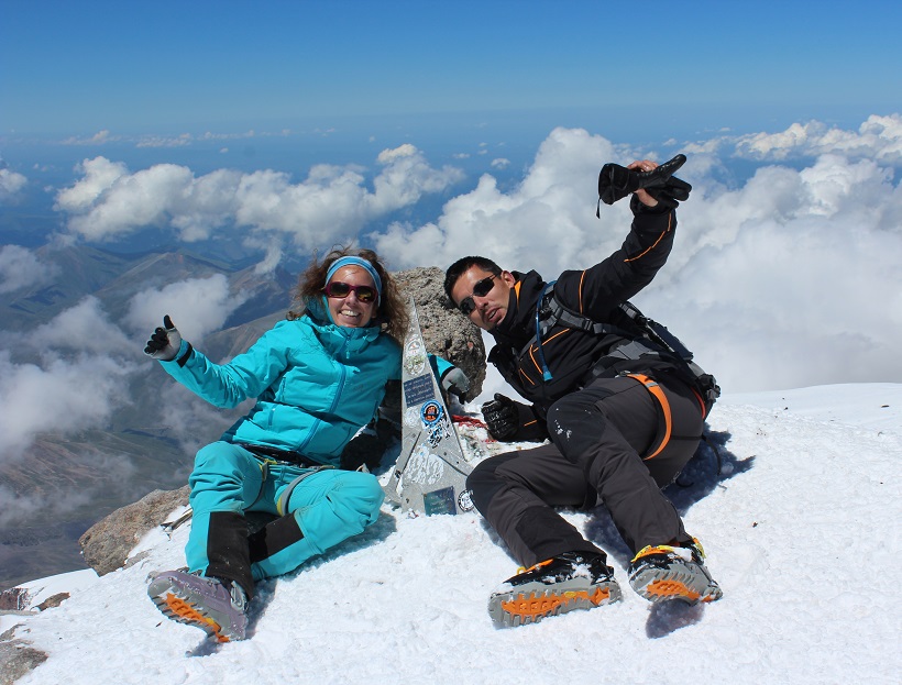 Voyage au Sommet de l'Elbrouz (5642m). Une femme souriante et un homme allongés sur la neige font signe à la caméra.